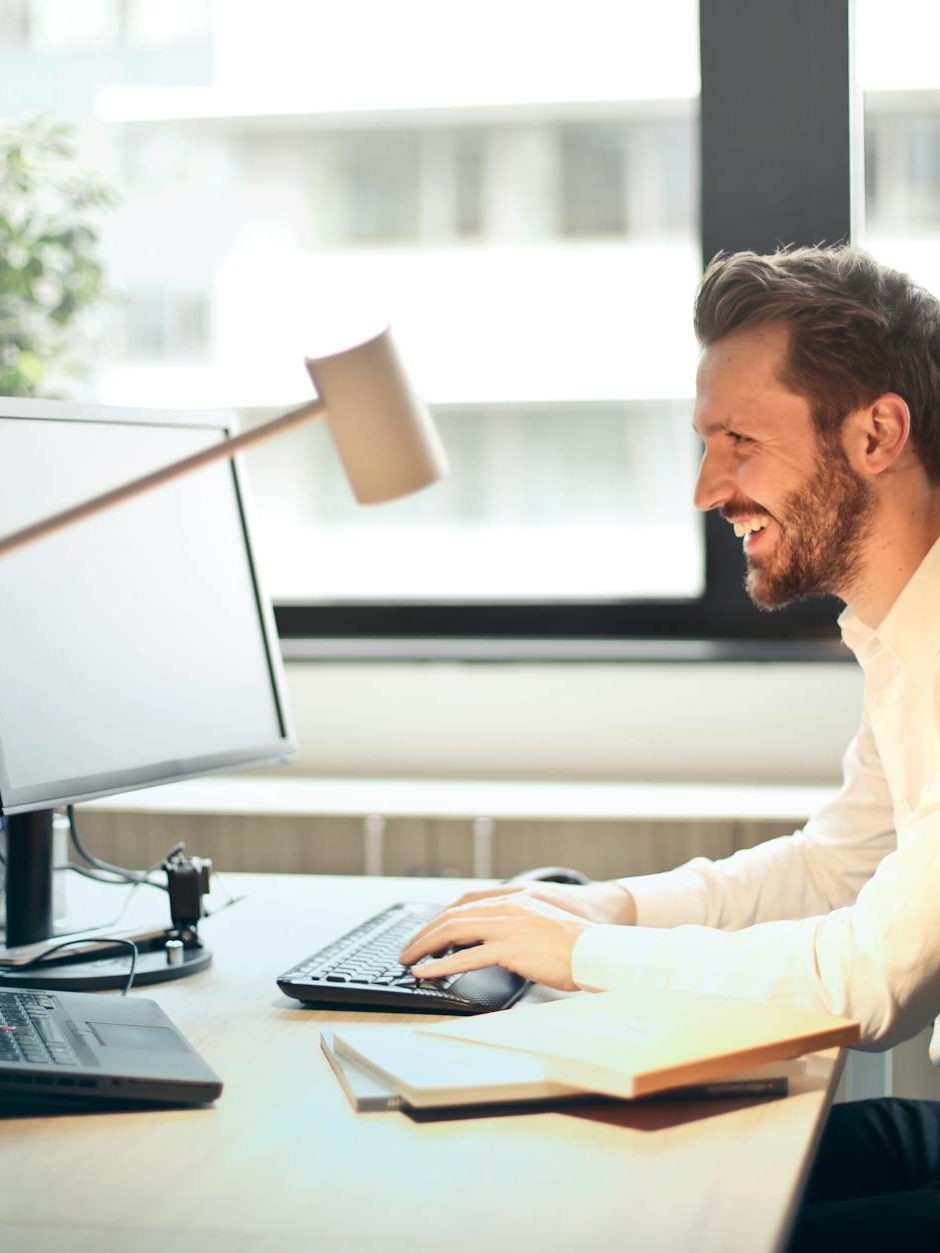 man in white dress shirt sitting on black rolling chair while facing black computer set and smiling
